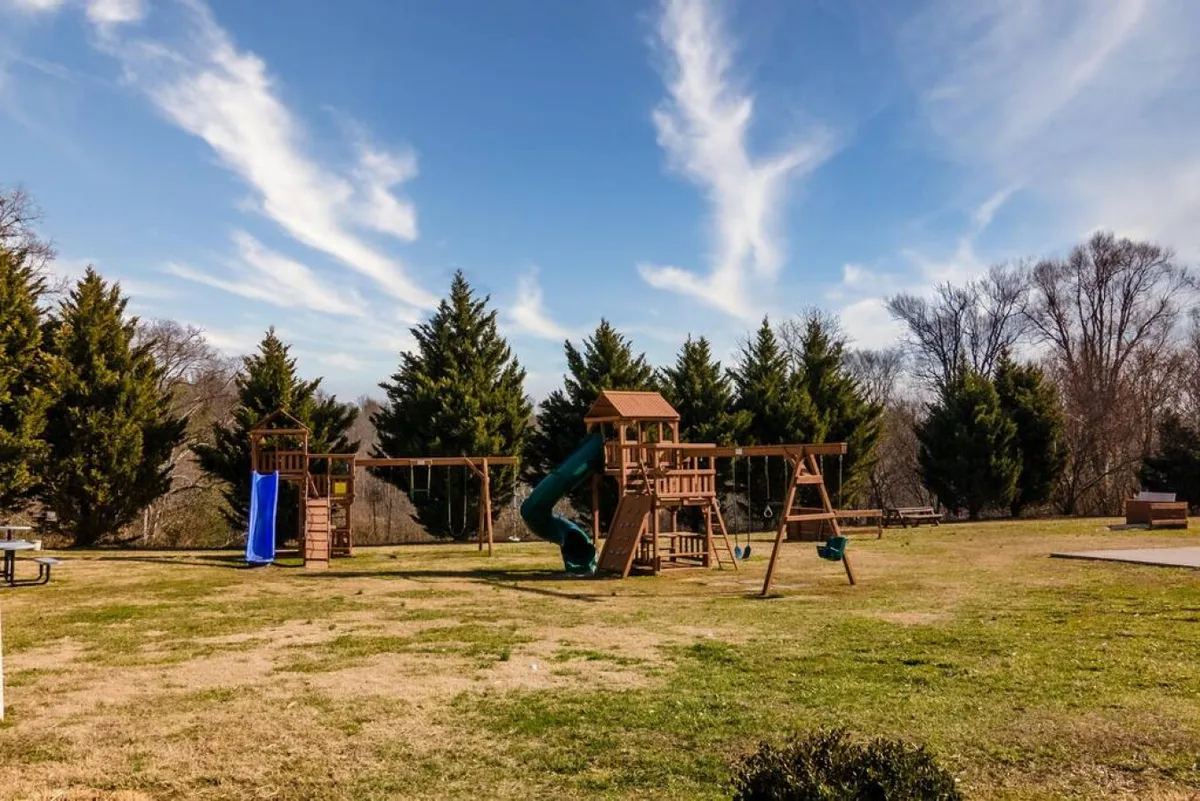 A cheerful playground scene with colorful equipment including swings and a slide, set next to a picnic area amidst green grass and trees. Children are playing, and families gather for outdoor fun, reflecting an atmosphere of joy and activity.