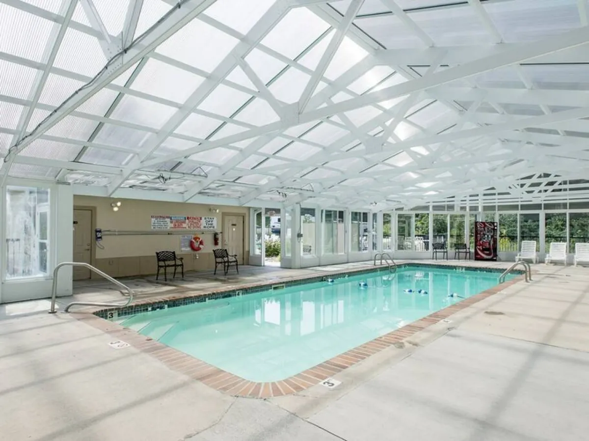 A bright, inviting indoor pool area featuring a large swimming pool under white ceiling beams. Comfortable seating lines the poolside, creating a relaxing environment for guests to unwind or socialize. The space is airy, well-lit, and exudes a refreshing, resort-like vibe.