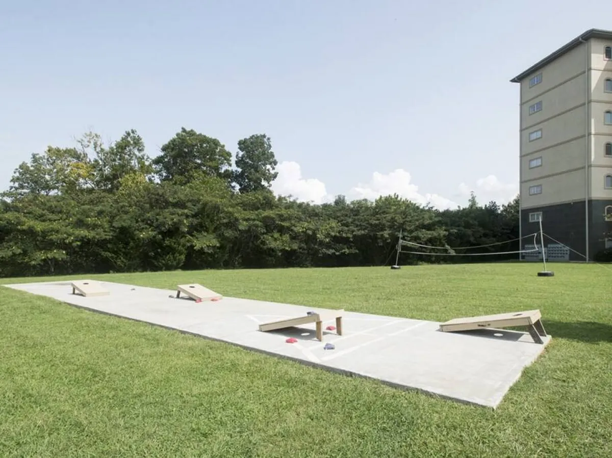 A green grassy outdoor space with cornhole boards set up, distant trees, and a building in the background. The scene is playful and casual, suggesting opportunities for games and outdoor gatherings.