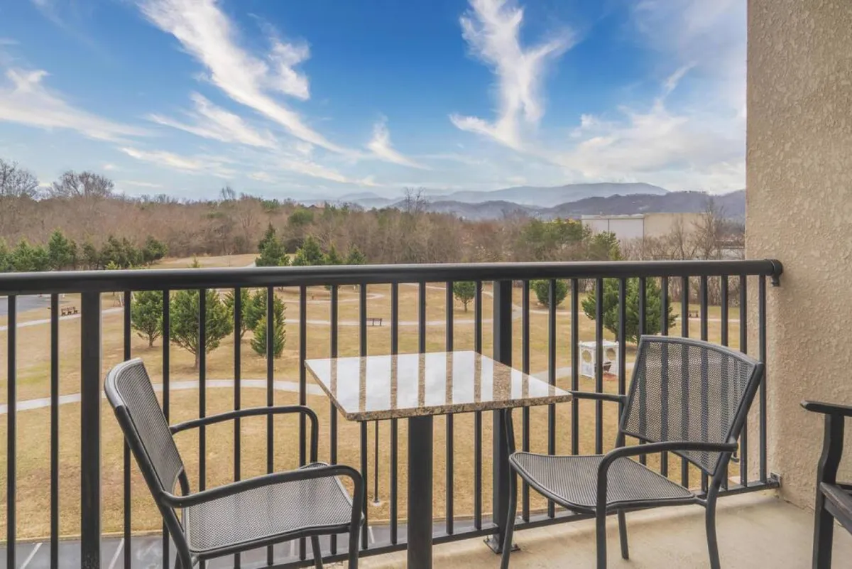 A private balcony with comfortable seating overlooks a sweeping mountain landscape and a serene open grassy area, inviting guests to relax and enjoy the outdoors. The setting is peaceful, with hints of morning dew on the grass and vibrant sunsets visible in the distance.
