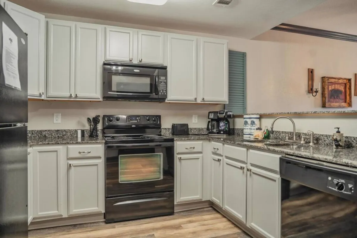 A bright, inviting kitchen scene displaying granite countertops, black appliances, white cabinetry, and a dedicated coffee station. The setup is tailored for easy meal preparation and group gatherings, with a cozy yet functional atmosphere.