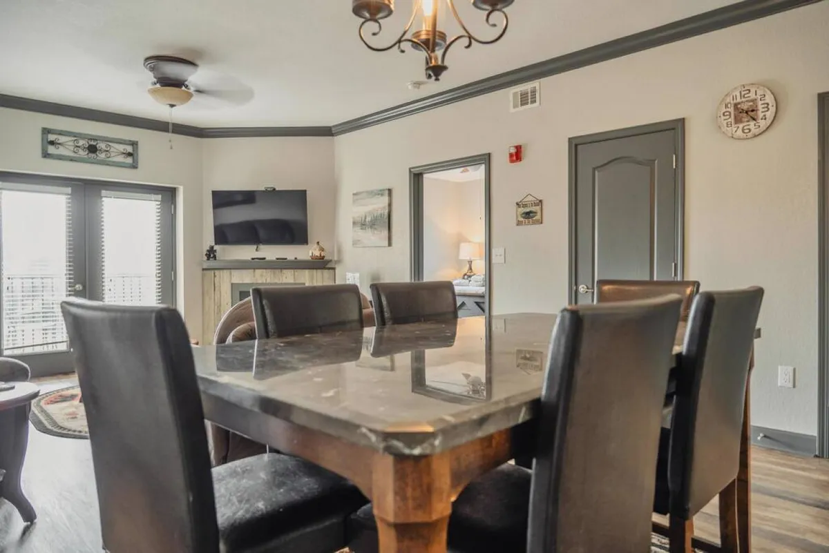 A dining area with a welcoming dining table, a stylish chandelier, decorative wall art, and a television visible in the background. The arrangement suggests space for meals, games, and shared memories, with easy access to adjoining rooms.