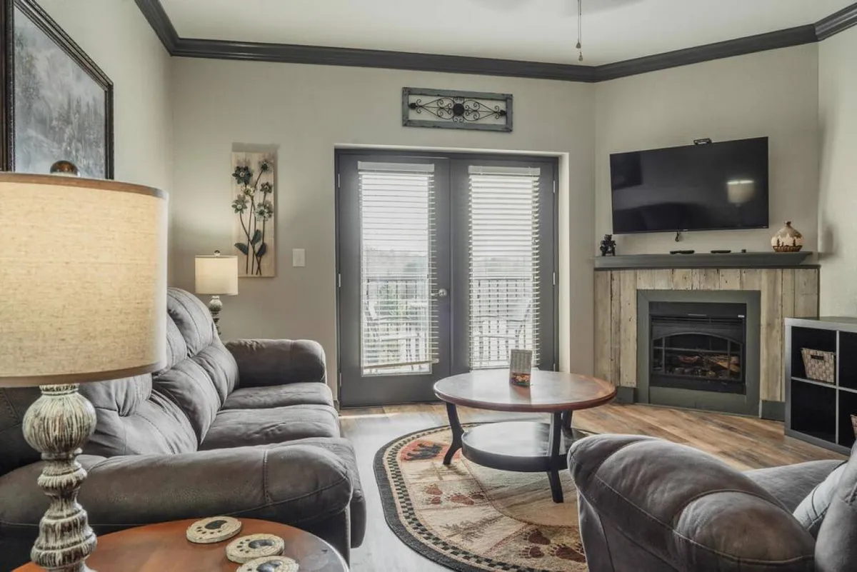 A bright and spacious living room featuring a plush sectional sofa, coffee table, fireplace, and a large flat-screen television. Double doors lead outside, allowing natural light to fill the space. The room is styled for comfort and group gatherings, with inviting decor and a relaxed atmosphere.