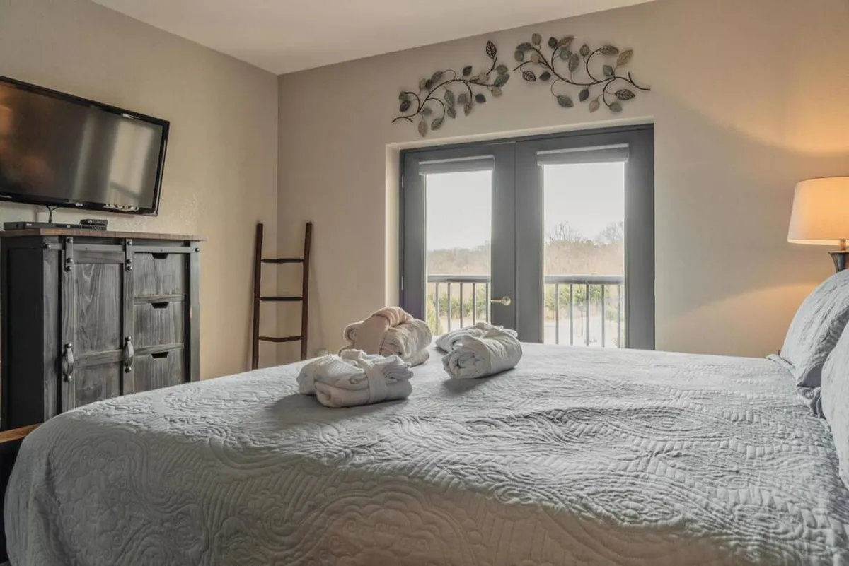 A bright, comfortable bedroom scene with a neatly arranged bed, decorative wall art, and a window allowing natural light to filter in. Towels are thoughtfully placed, and the space feels airy and peaceful.